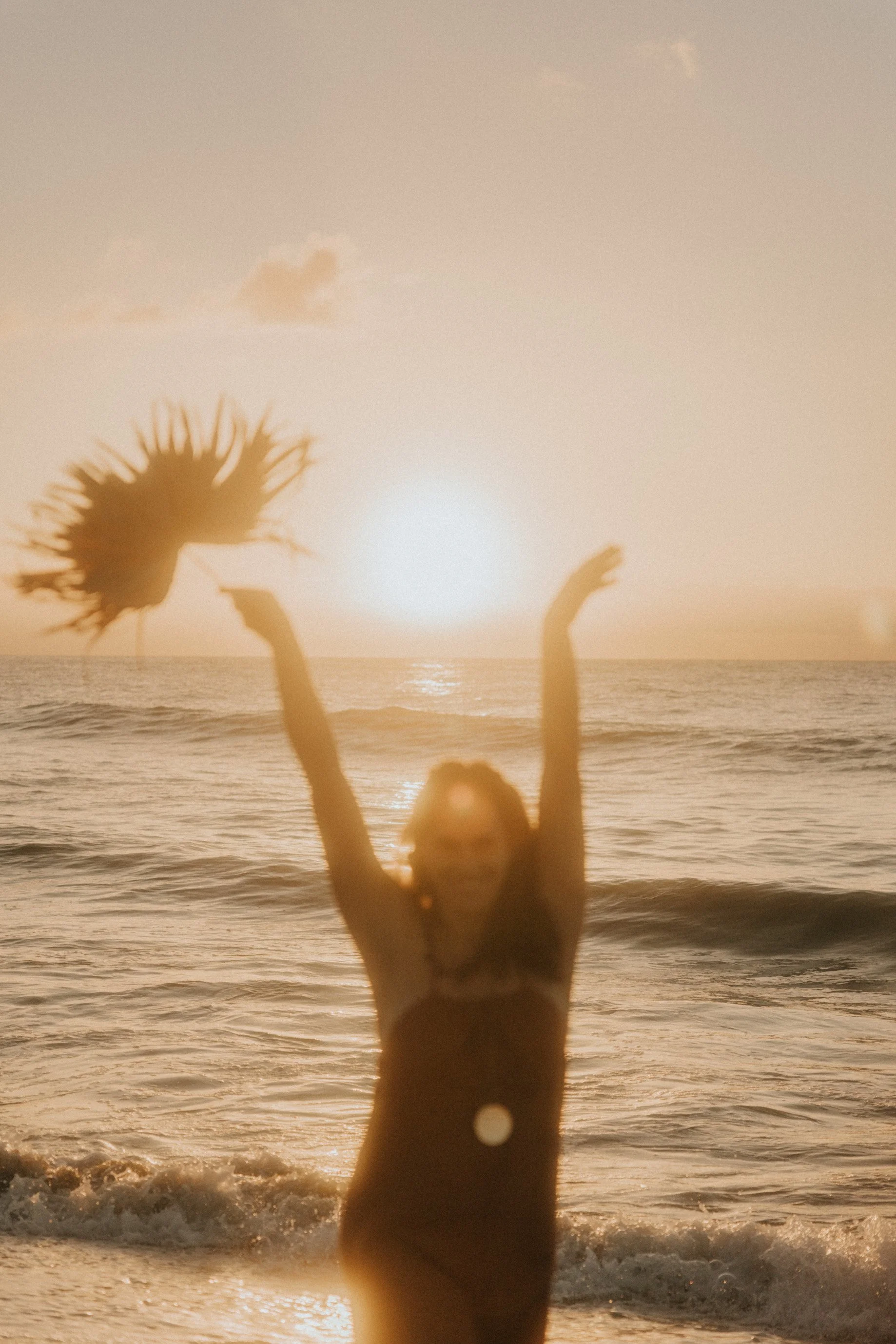 Mujer en la playa durante el atardecer, levantando los brazos con una sonrisa, sosteniendo un sombrero, con el sol en el horizonte y el océano de fondo.