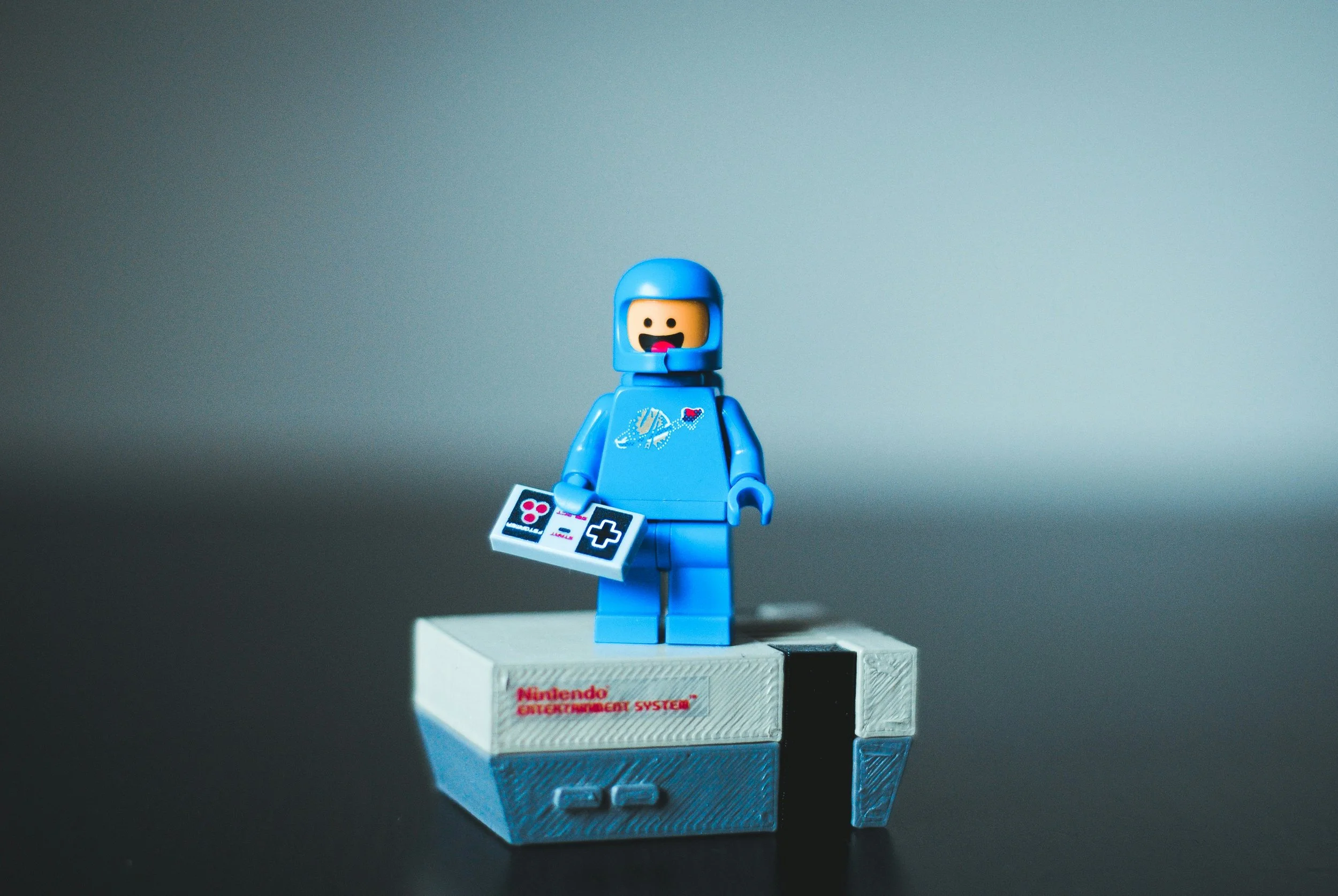 Lego figurine of a person in a blue space suit holding a controller, standing on a Nintendo Entertainment System console