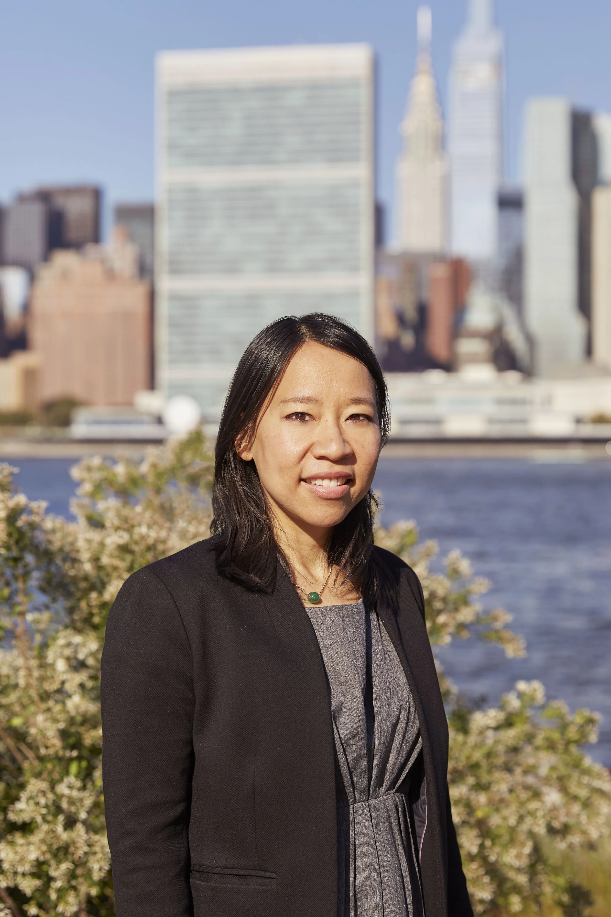A woman standing outdoors in front of a city skyline, including the Empire State Building, with water and flowering bushes in the background.