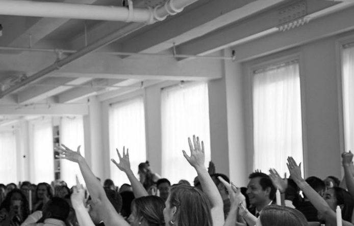 A group of people participating in a lively indoor event, raising their hands with some smiling, in a room with white walls and large windows.