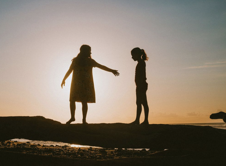 Two silhouetted women on a sandy beach at sunset, one reaching out to the other.