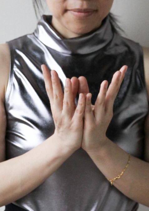 A young girl in a shiny black and white sleeveless top with her hands pressed together in a prayer or greeting gesture.