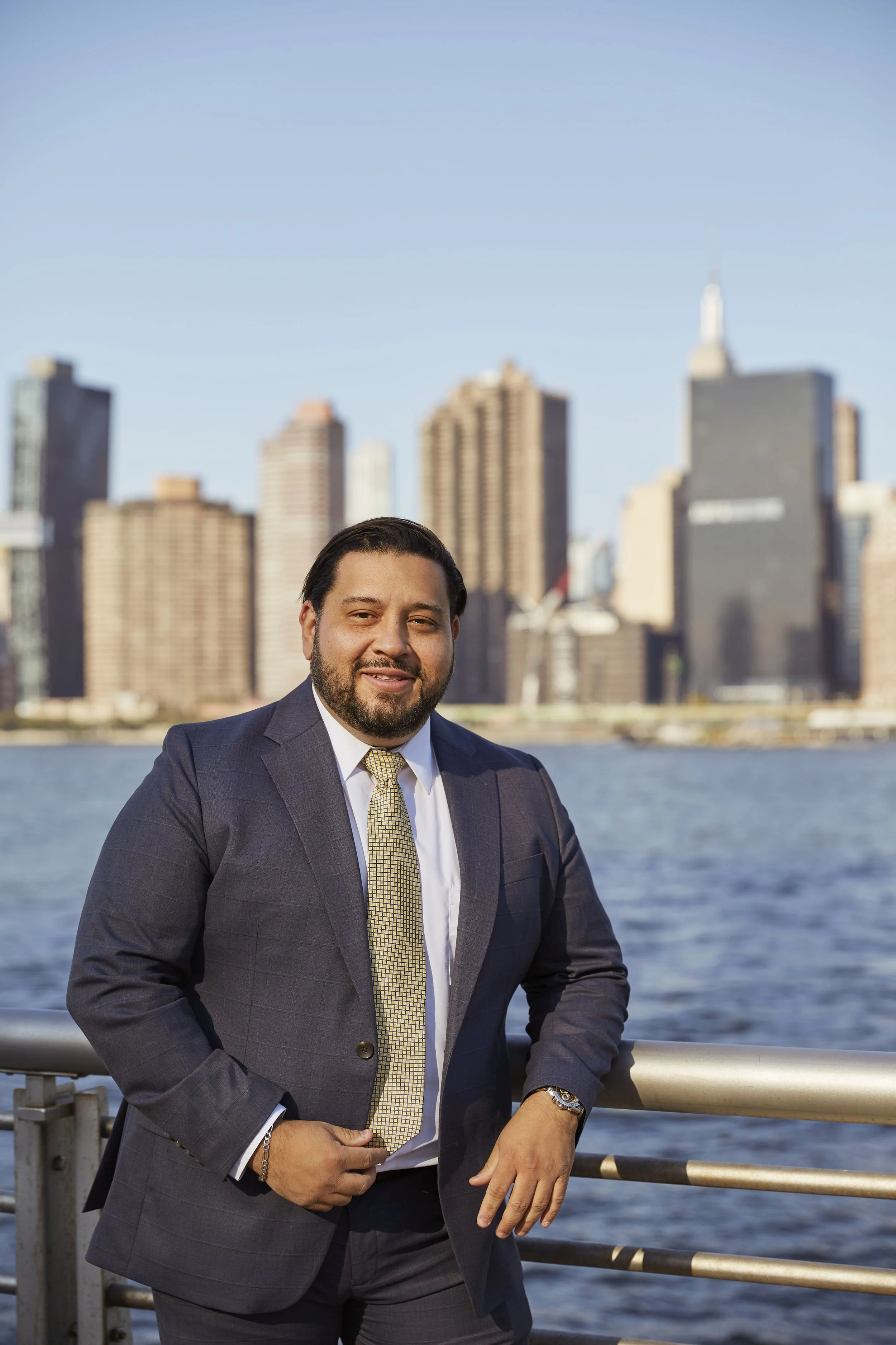 A man in a suit standing by the water with a city skyline in the background.