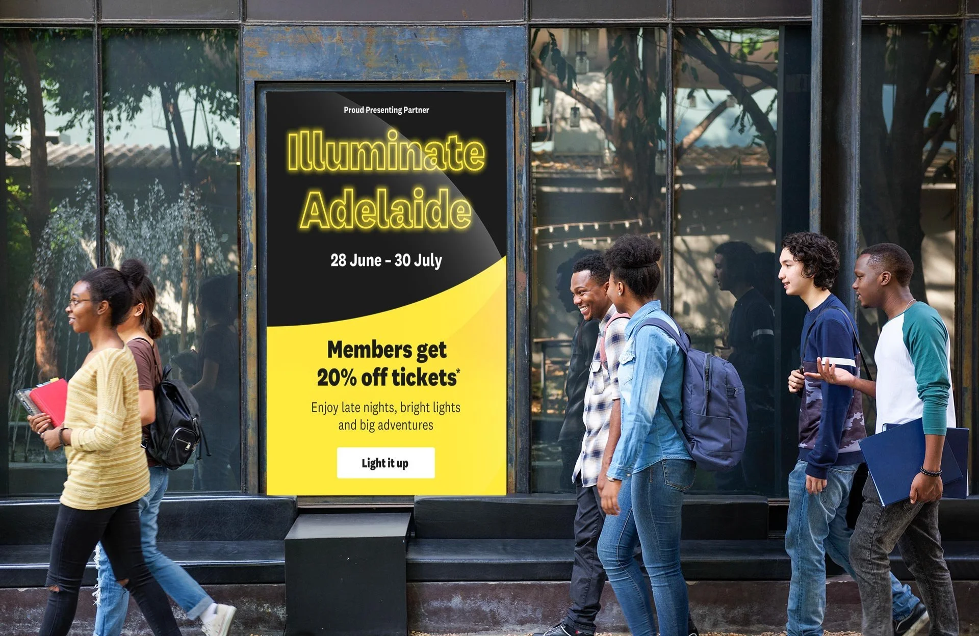 A group of diverse young adults walking past a large glass window with an illuminated poster advertising the Illuminate Adelaide event, which runs from June 28 to July 30, offering members 20% off tickets and featuring bright lights and adventures.
