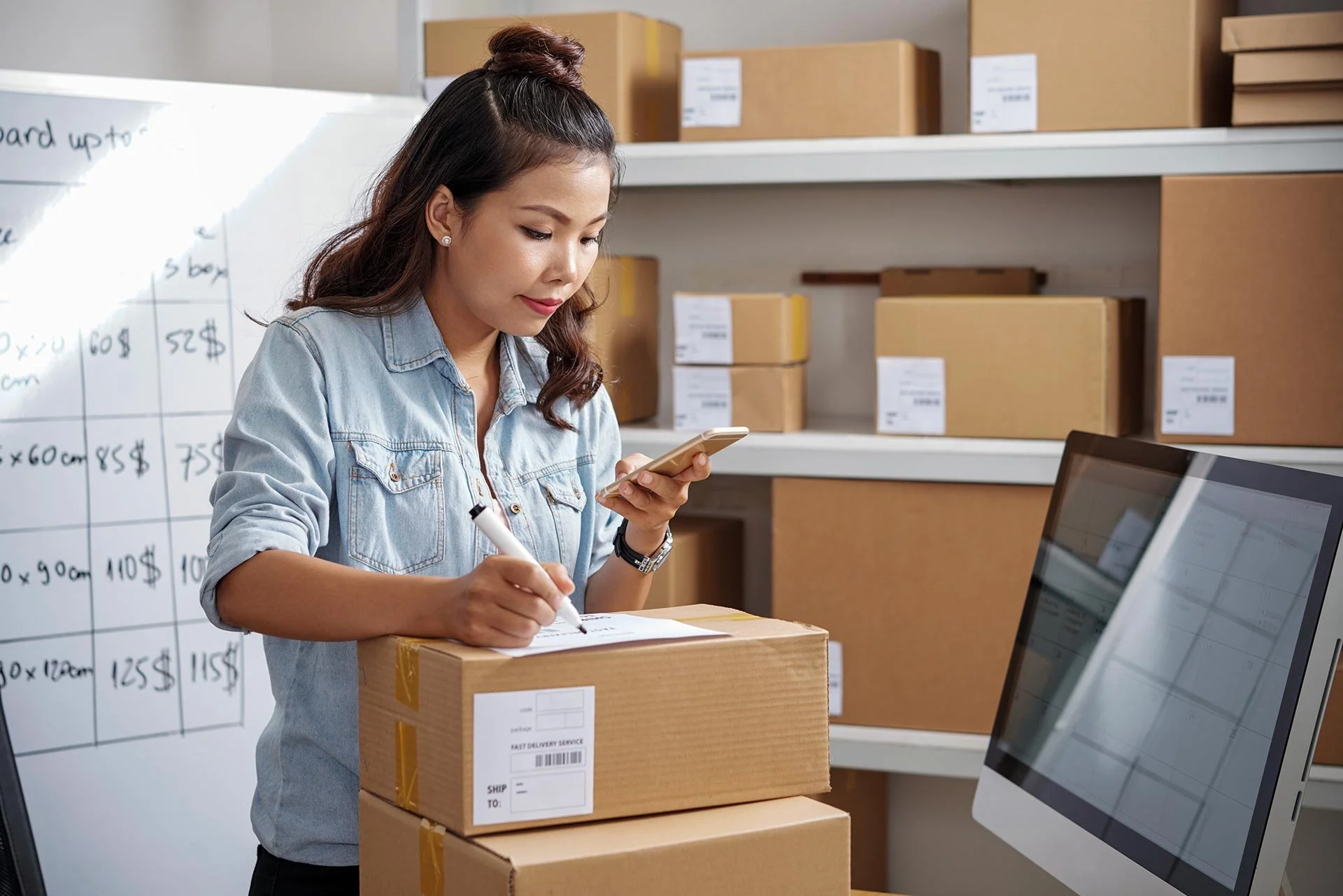 Woman working in a warehouse, holding a smartphone and marking a box with a marker.