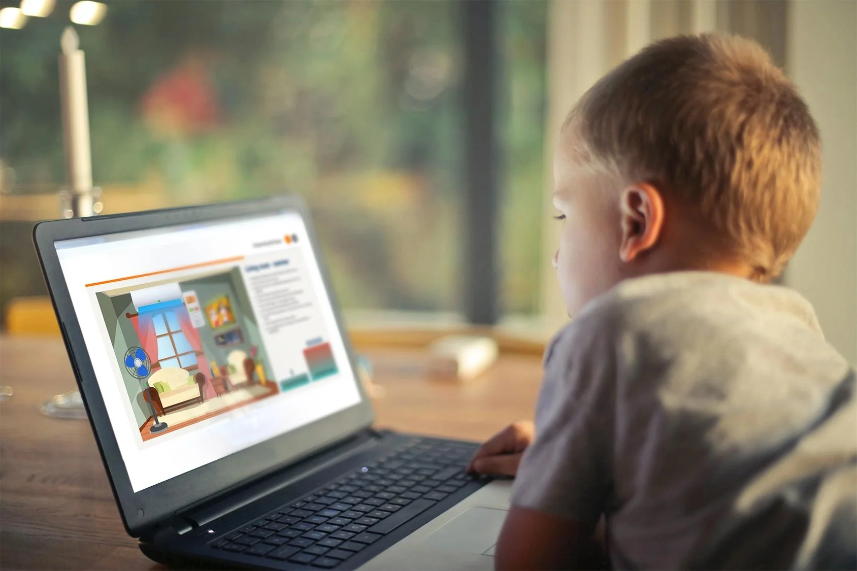 A young boy sitting at a wooden table, using a laptop to view an online interior design or home improvement website with a colorful room illustration.
