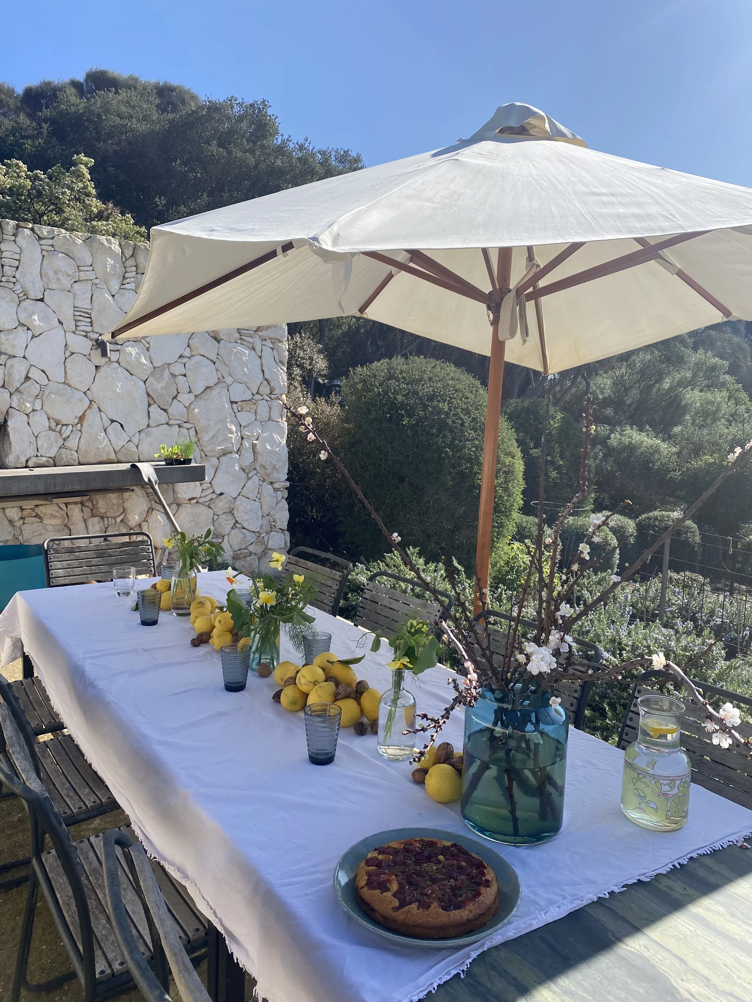 Outdoor dining table with white tablecloth, decorated with yellow fruits, vases with flowers, and set glasses under a large white umbrella. Background features a stone wall, green bushes, and trees with a clear blue sky.