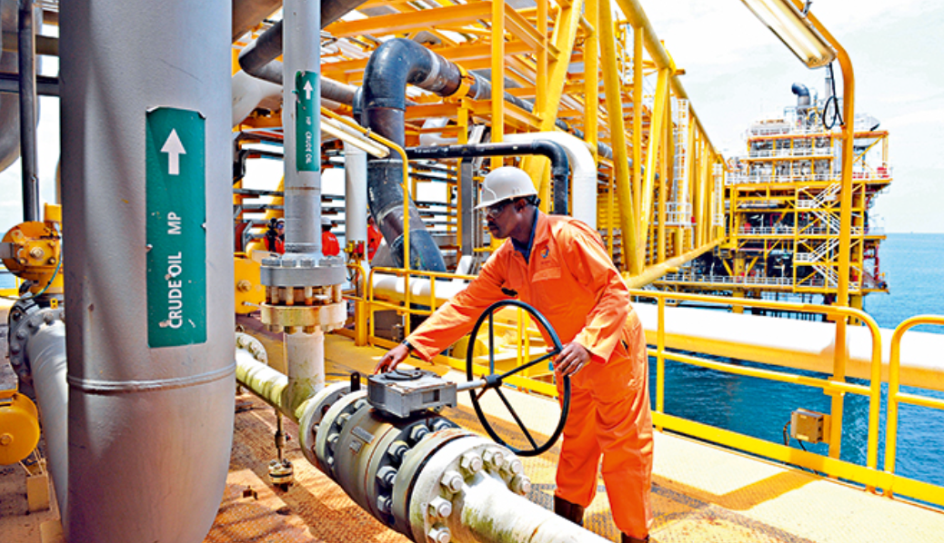 A worker in orange coveralls and a white helmet operating a valve on an offshore oil platform with piping and metal structures.