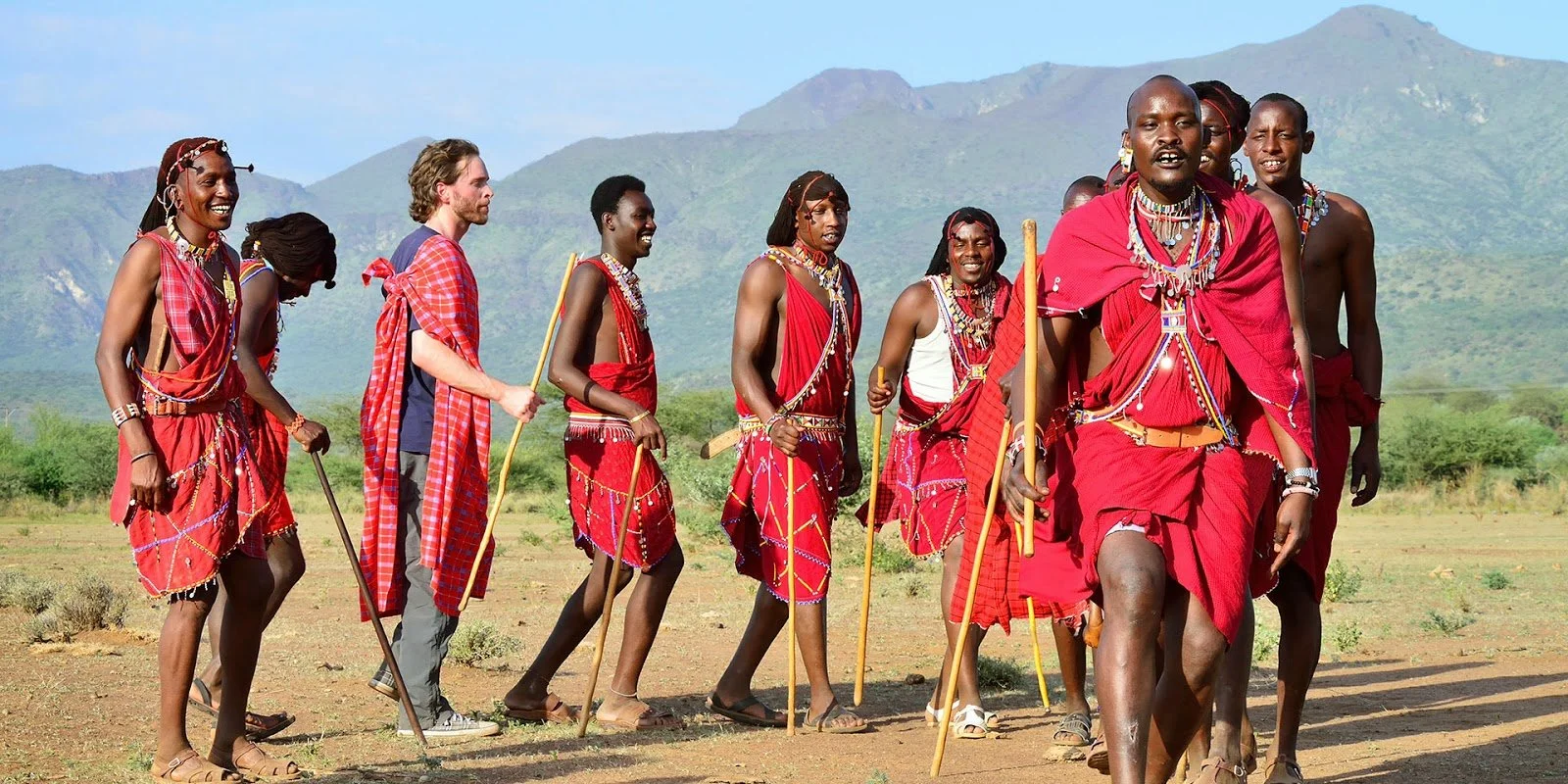 Group of Maasai men and boys in traditional red shúkā outfits, walking outdoors with mountains in the background.