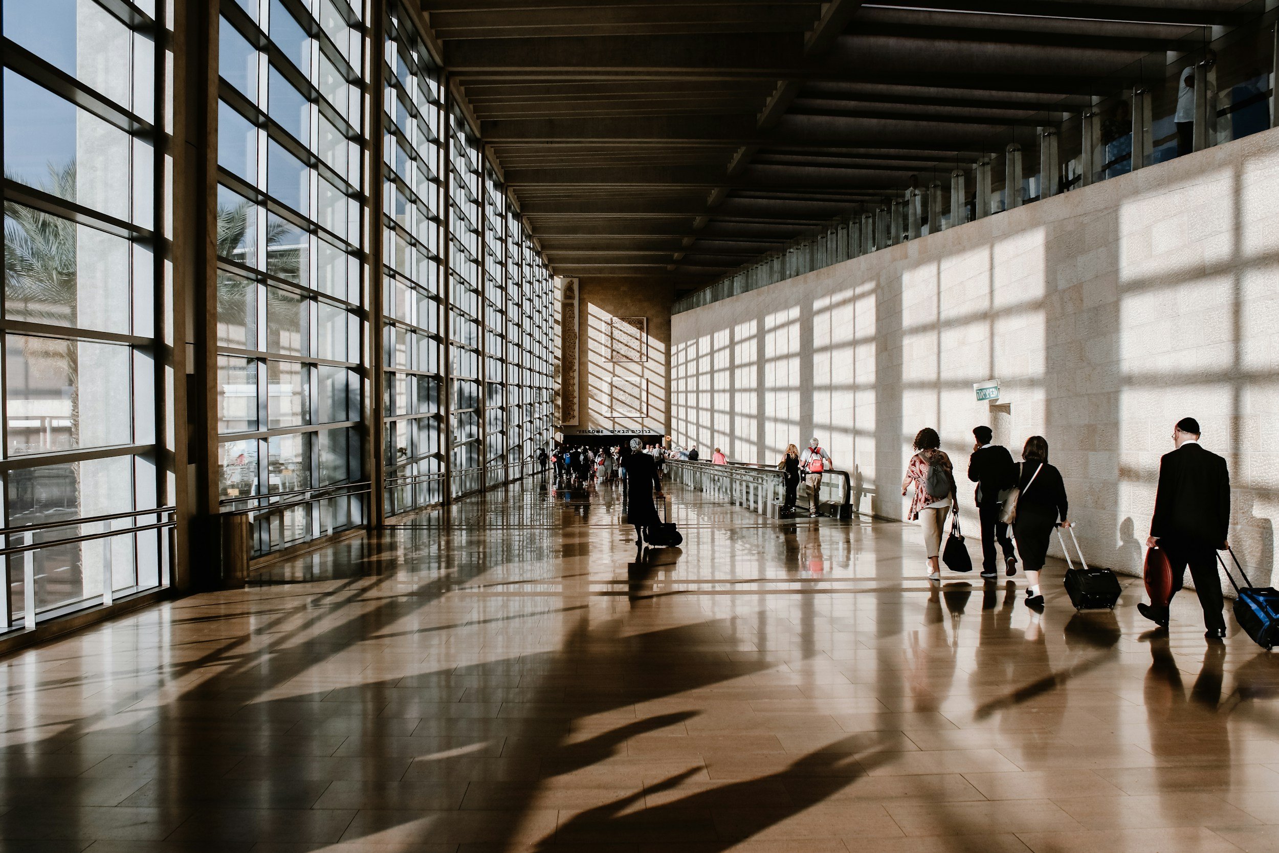 People walking inside an airport terminal with large glass windows and sunlight casting shadows on the floor.