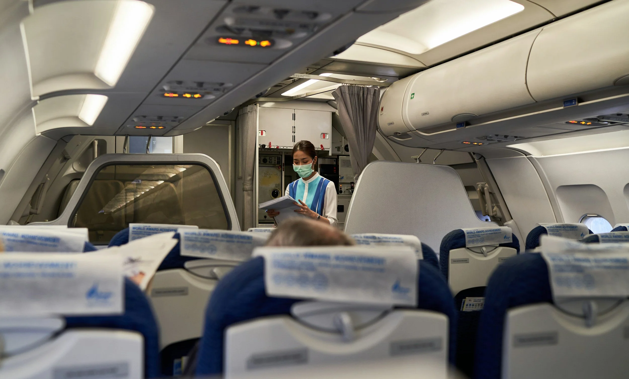 A flight attendant wearing a face mask and uniform reading a document on an airplane.