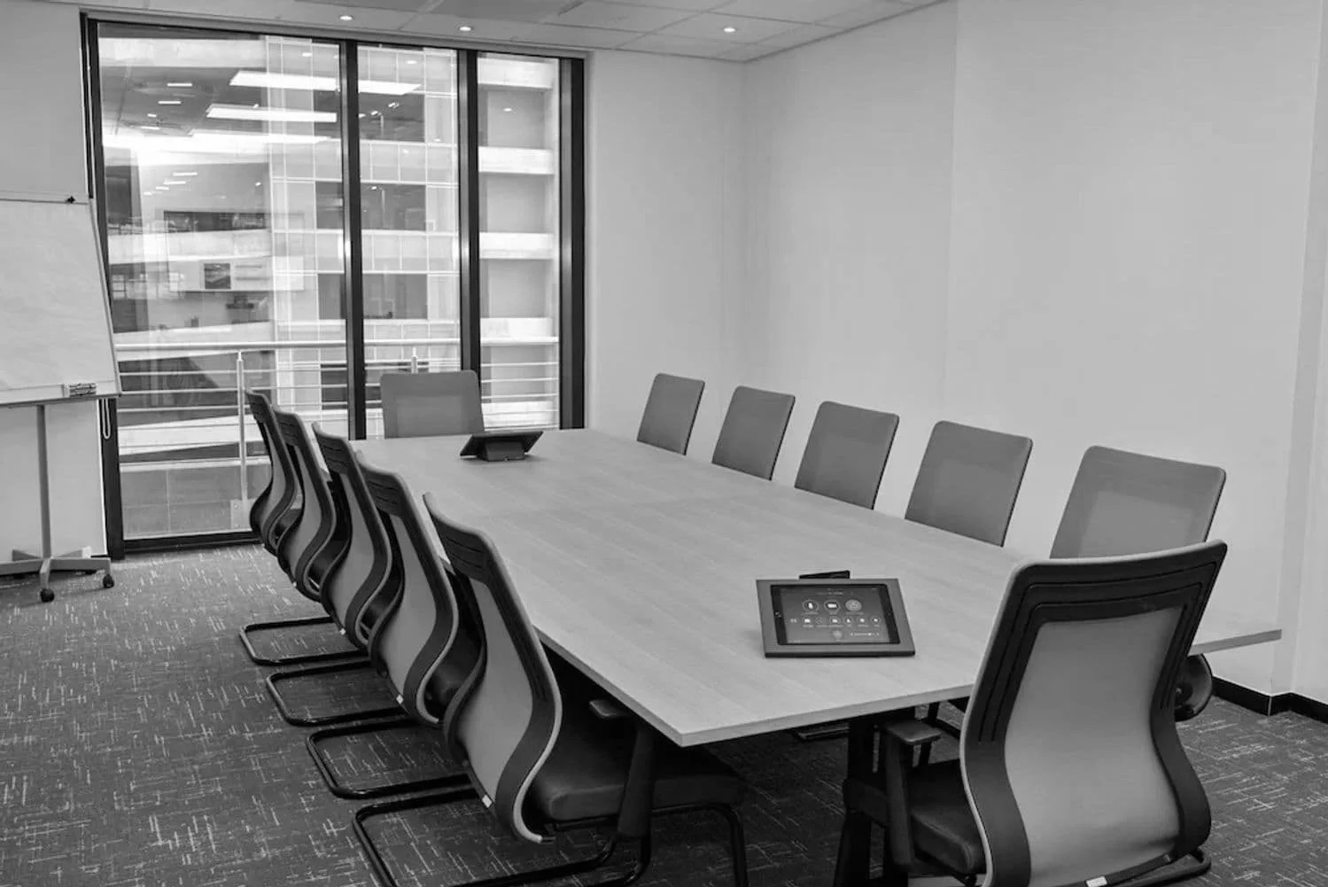 Empty modern conference room with a long table, ten chairs, a tablet device on the table, a whiteboard on a stand, and large windows revealing a building exterior.