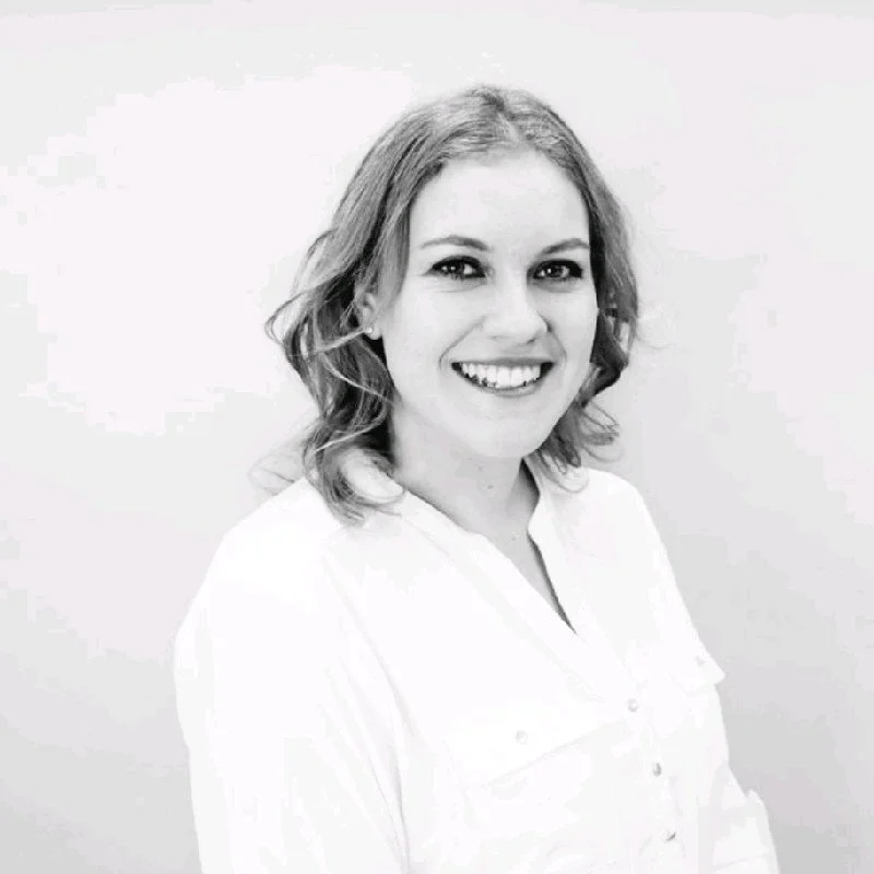 Black and white portrait of a woman smiling, with shoulder-length wavy hair, wearing a white shirt, standing against a plain white background.