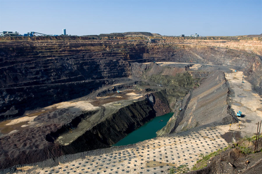 Open-pit copper mining quarry with terraced excavation walls and a central water-filled pit, surrounded by construction equipment.