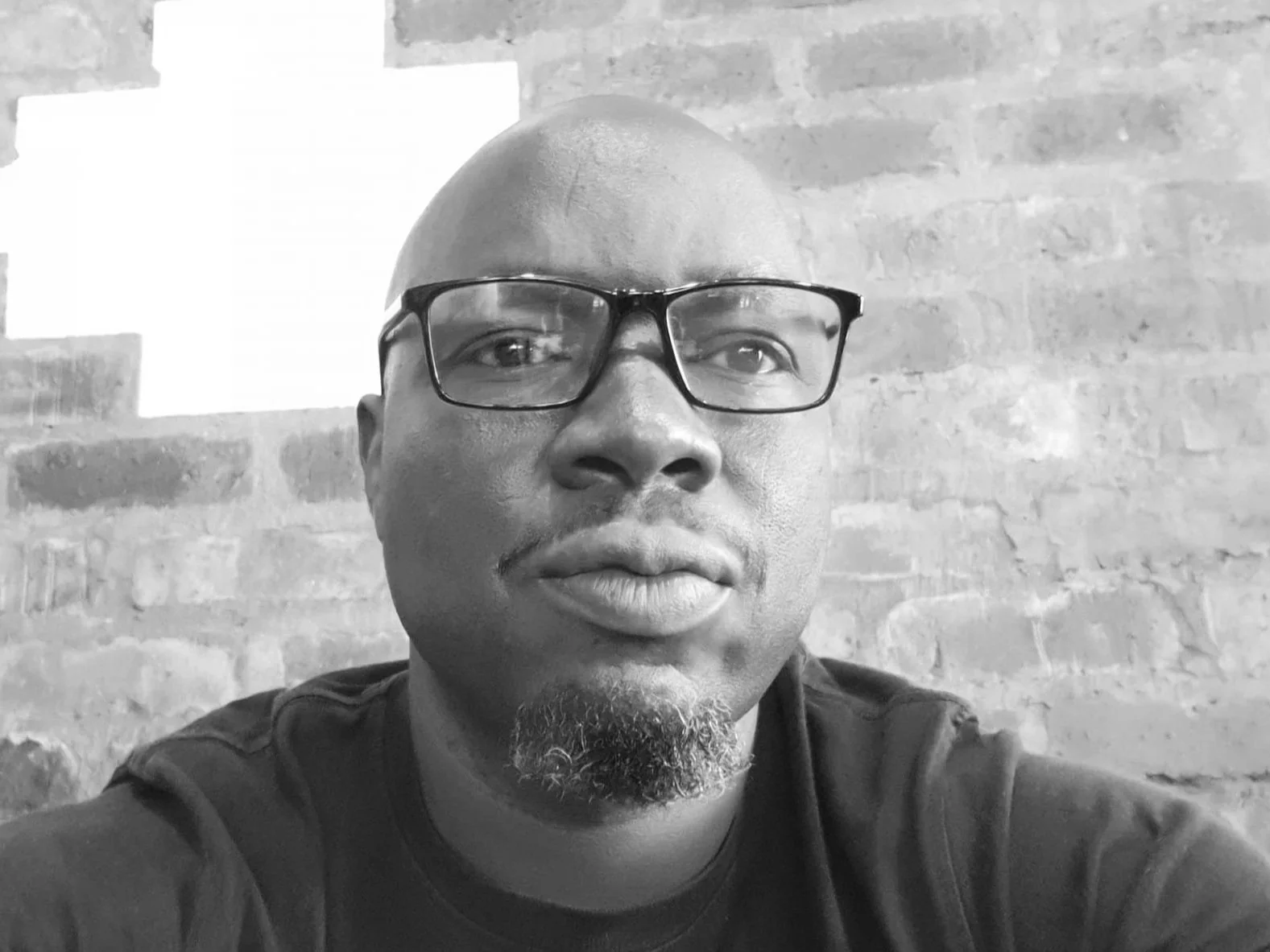 A black-and-white photo of a happy customer who is man wearing glasses, with a bald head and a goatee, posing in front of a brick wall.