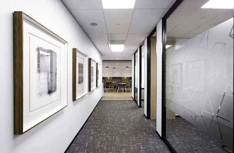 Modern office hallway with framed artwork on the left wall and glass-walled conference rooms on the right, leading to a meeting area with chairs and pendant lighting.