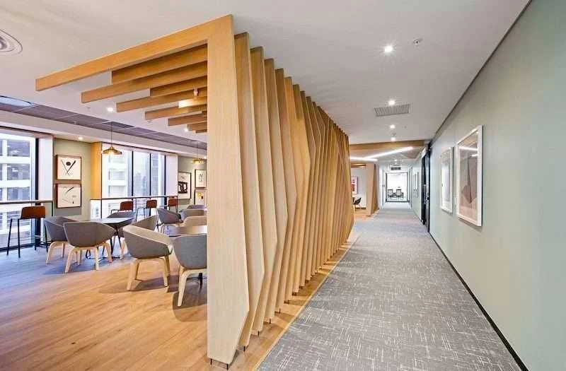 Modern interior of a building with a wooden partition separating a lounge area with chairs and tables from a hallway. The lounge has large windows, framed artwork, and contemporary lighting.