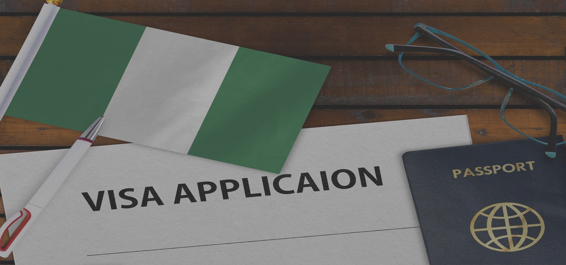 A printed visa application form, a small Mexican flag, a passport with a globe logo, a pair of eyeglasses, and a pen on a wooden surface.
