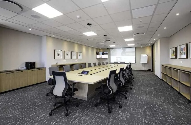 A modern conference room with a large U-shaped table, ergonomic office chairs, and a projection screen at the front. The room has framed artwork on the walls, a side cabinet with refreshments, and built-in shelving.