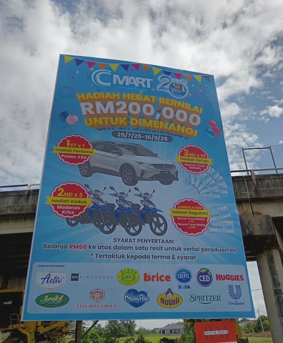 A promotional billboard for a contest at a store called C-Mart celebrating its 20th anniversary. The billboard details prizes including a car, motorcycles, cash vouchers, and a cash prize, with participation requirements of spending RM60 in a single receipt. The event runs from July 25 to September 16, 2025, and features sponsor logos at the bottom.