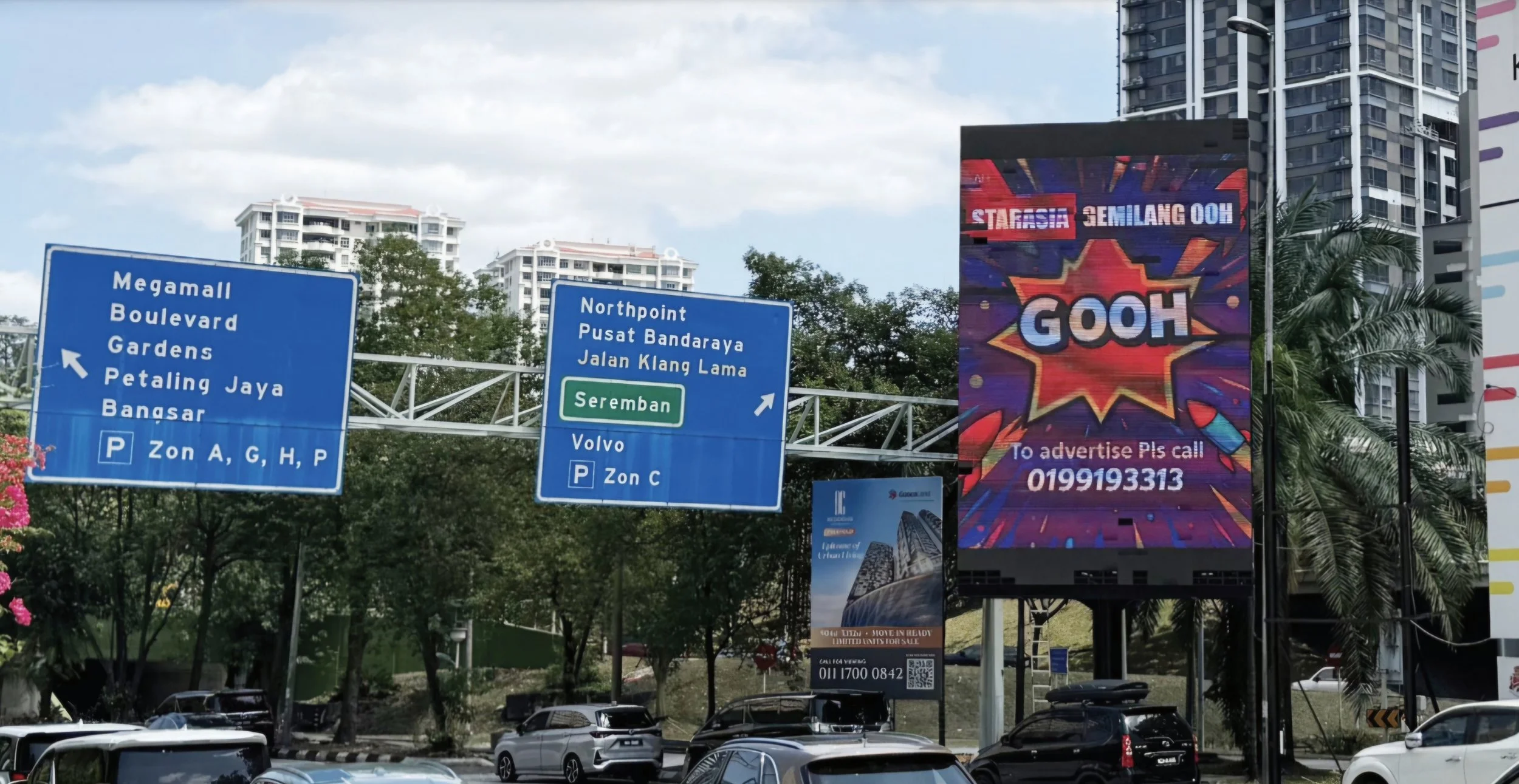 Urban street scene with blue directional signs and a large colorful electronic billboard displaying advertisements, with trees and high-rise buildings in the background and cars parked along the street near Lingakaran Syed Putra, MidValley KL.