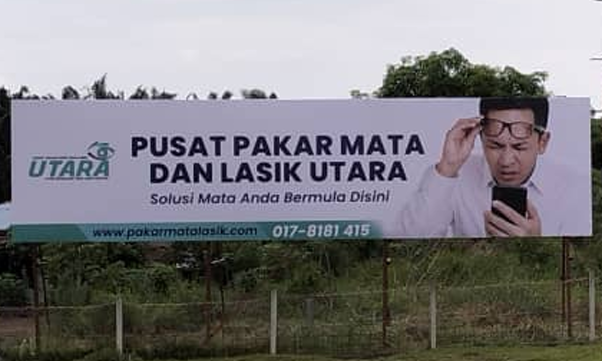Billboard advertising an eye care center called Pusat Pakar Mata dan Lasik Utara, with a man holding glasses and looking at his phone.