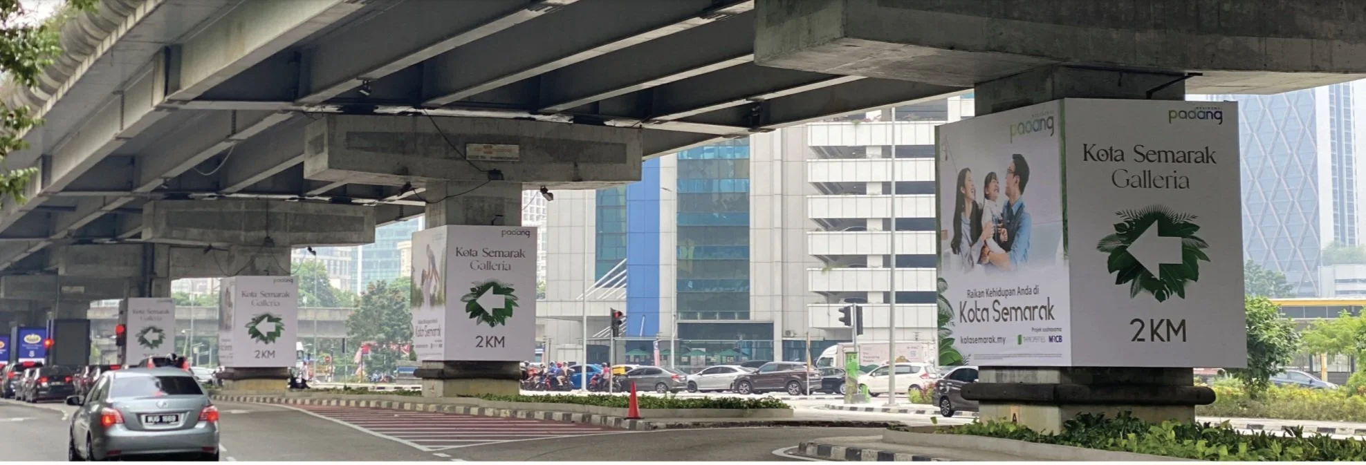 Urban street scene with large road signs indicating 2 kilometers to Kota Semarak Galleria, under an overpass. Buildings and traffic are visible in the background, and there are several cars on the road near Jalan Tun Razak, Kuala Lumpur.
