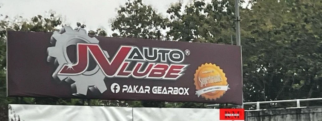Sign for JNAuto Lube with gear logo and Facebook page link, along with an orange badge or seal, outdoors with trees in the background.