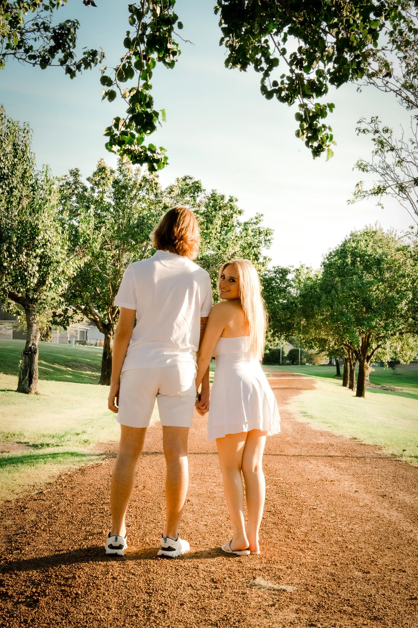 A couple holding hands and walking on a dirt path in a park, surrounded by green trees, under a light blue sky.
