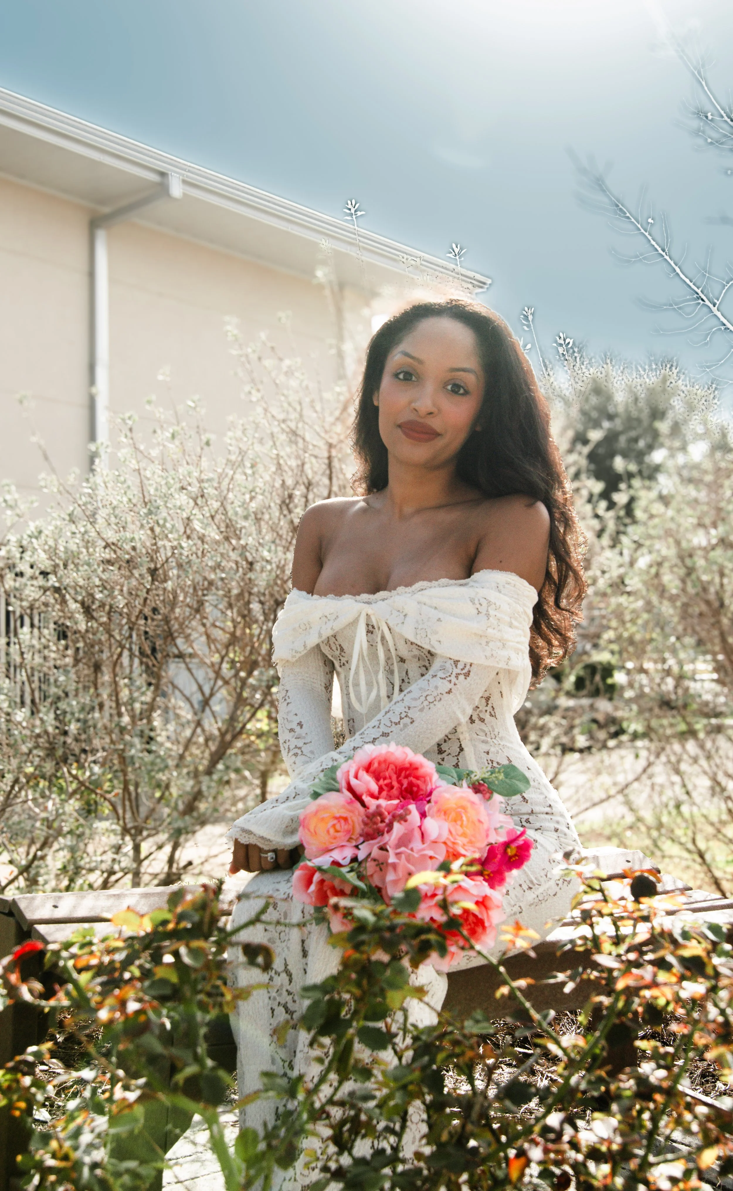 A woman with long dark hair in a white lace off-the-shoulder dress sitting on a park bench, holding a bouquet of pink and peach flowers, surrounded by blooming bushes and trees, with sunlight streaming from above.
