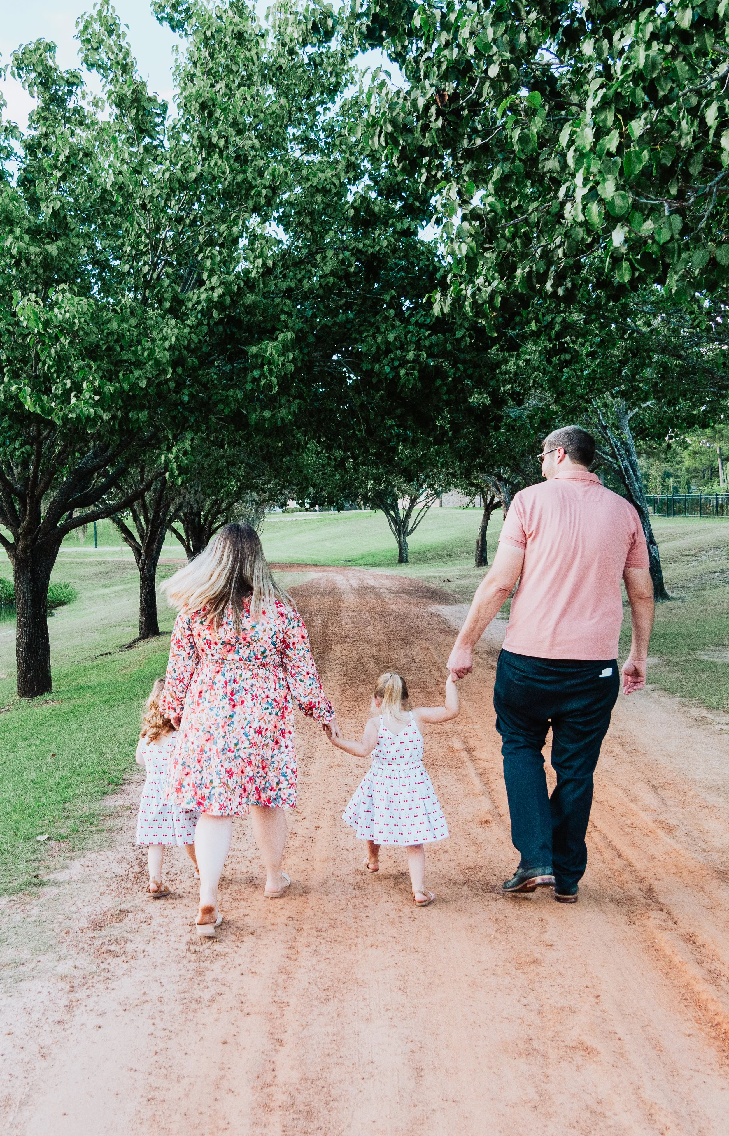 A family walking down a dirt path in a park, holding hands. The family includes two adults and three children, all dressed in casual clothing. Tall green trees line the path on both sides.