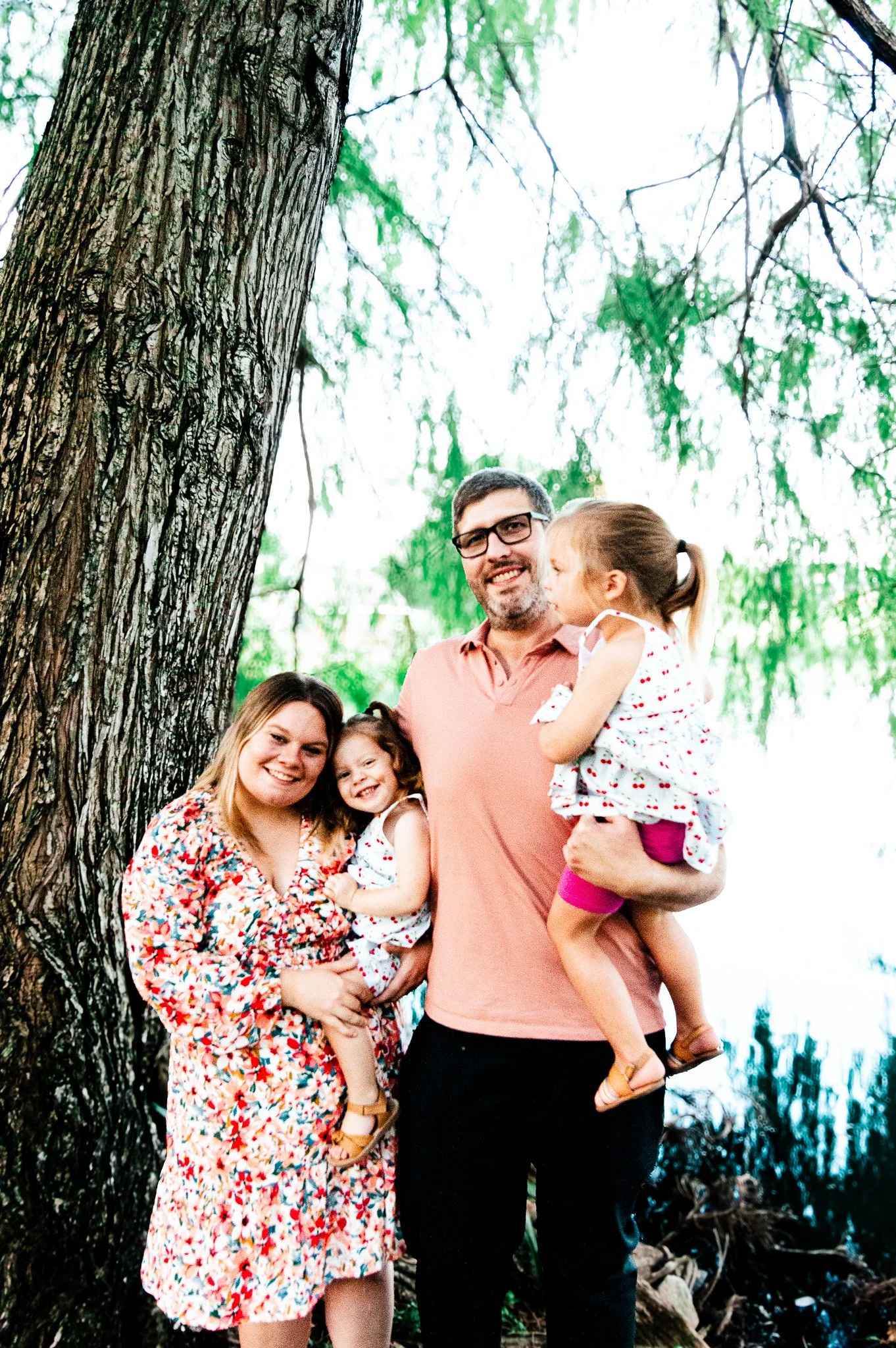 Family of four standing outdoors by a tree with green leaves, smiling. The mother holds a young girl, the father holds a young girl, and they are near a body of water.