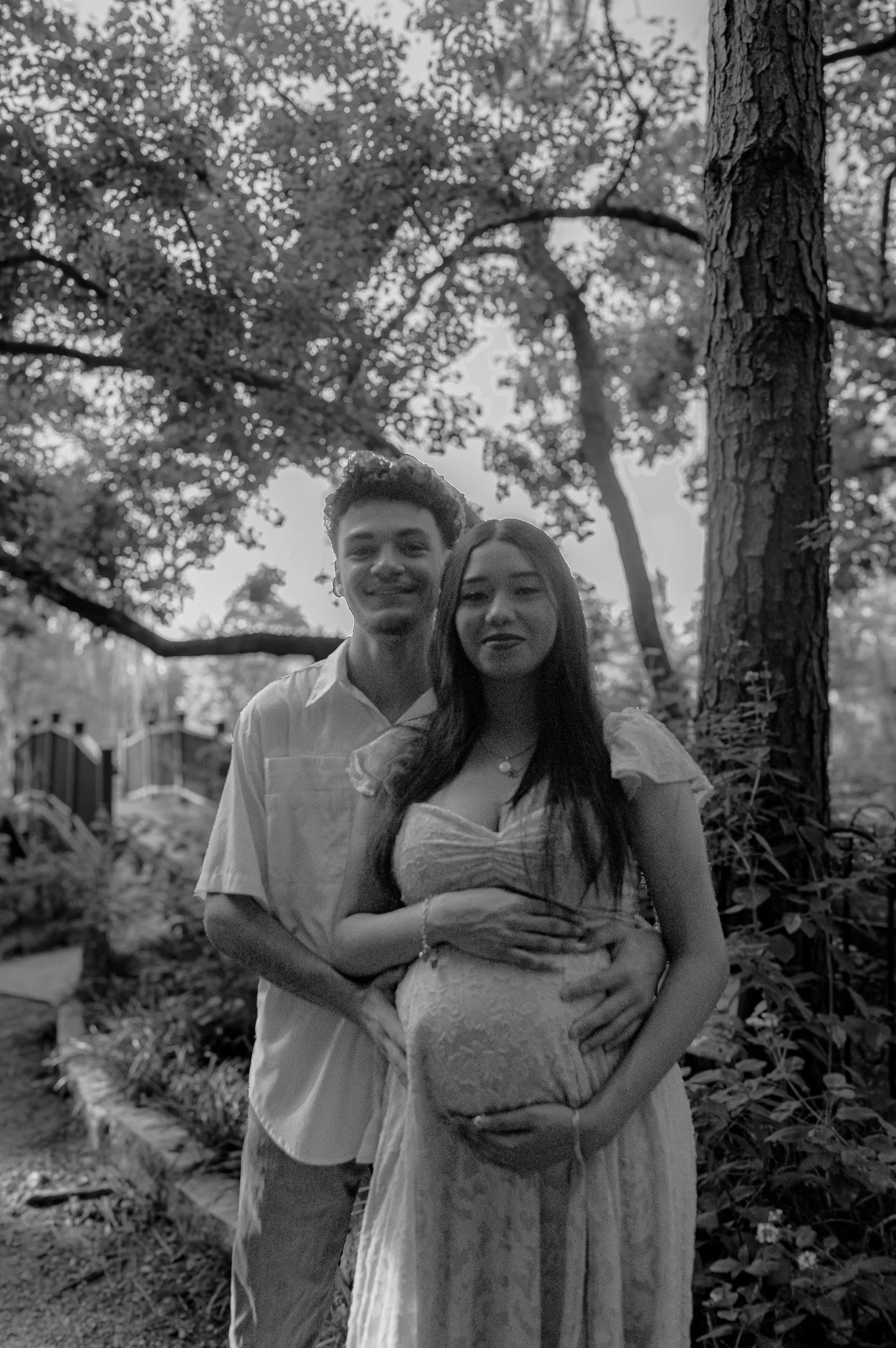 A black-and-white photo of a smiling couple, a man and a pregnant woman, standing outdoors in a wooded area with trees and a small bridge in the background.