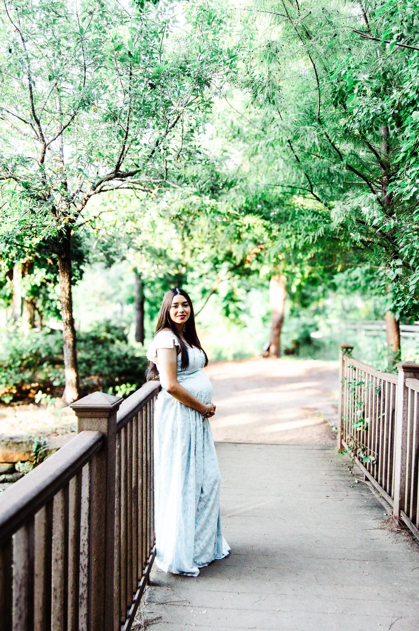 A pregnant woman standing on a wooden pathway in a lush green park, surrounded by trees and sunlight.