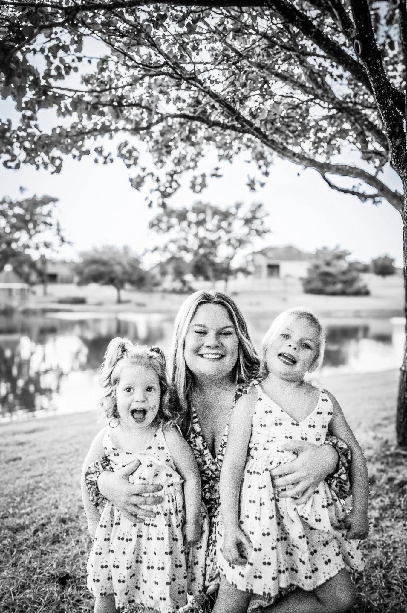A woman smiling and holding two young girls in a park near a pond, with trees and houses in the background, black and white photograph.