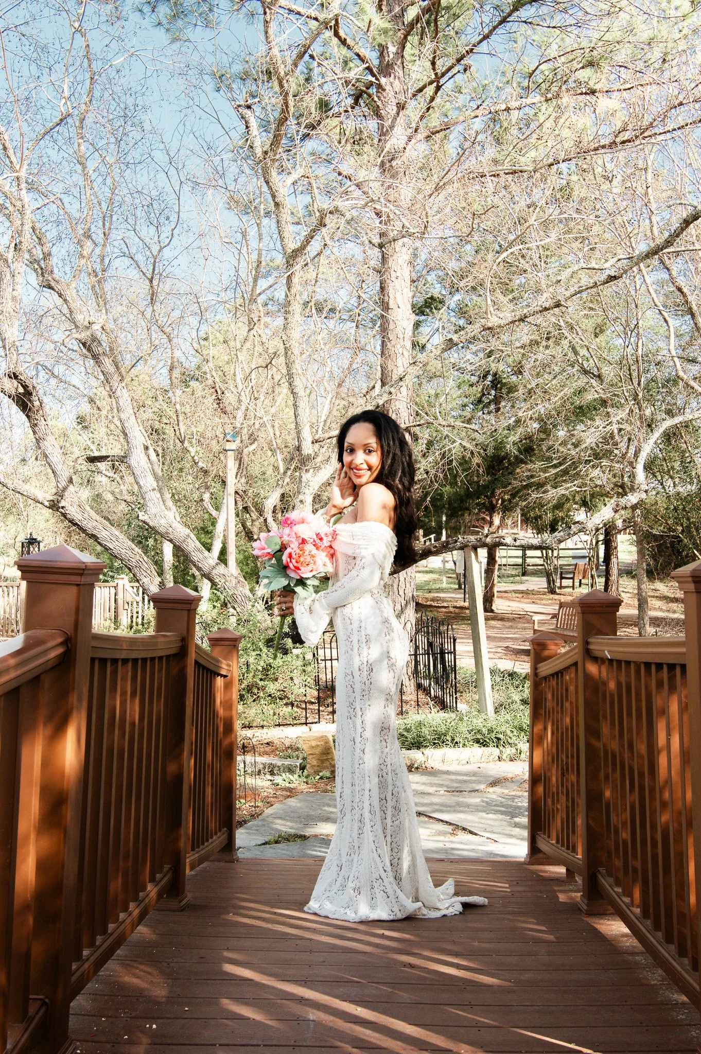 A woman in a white lace dress holding a bouquet of pink flowers standing on a wooden bridge outdoors.
