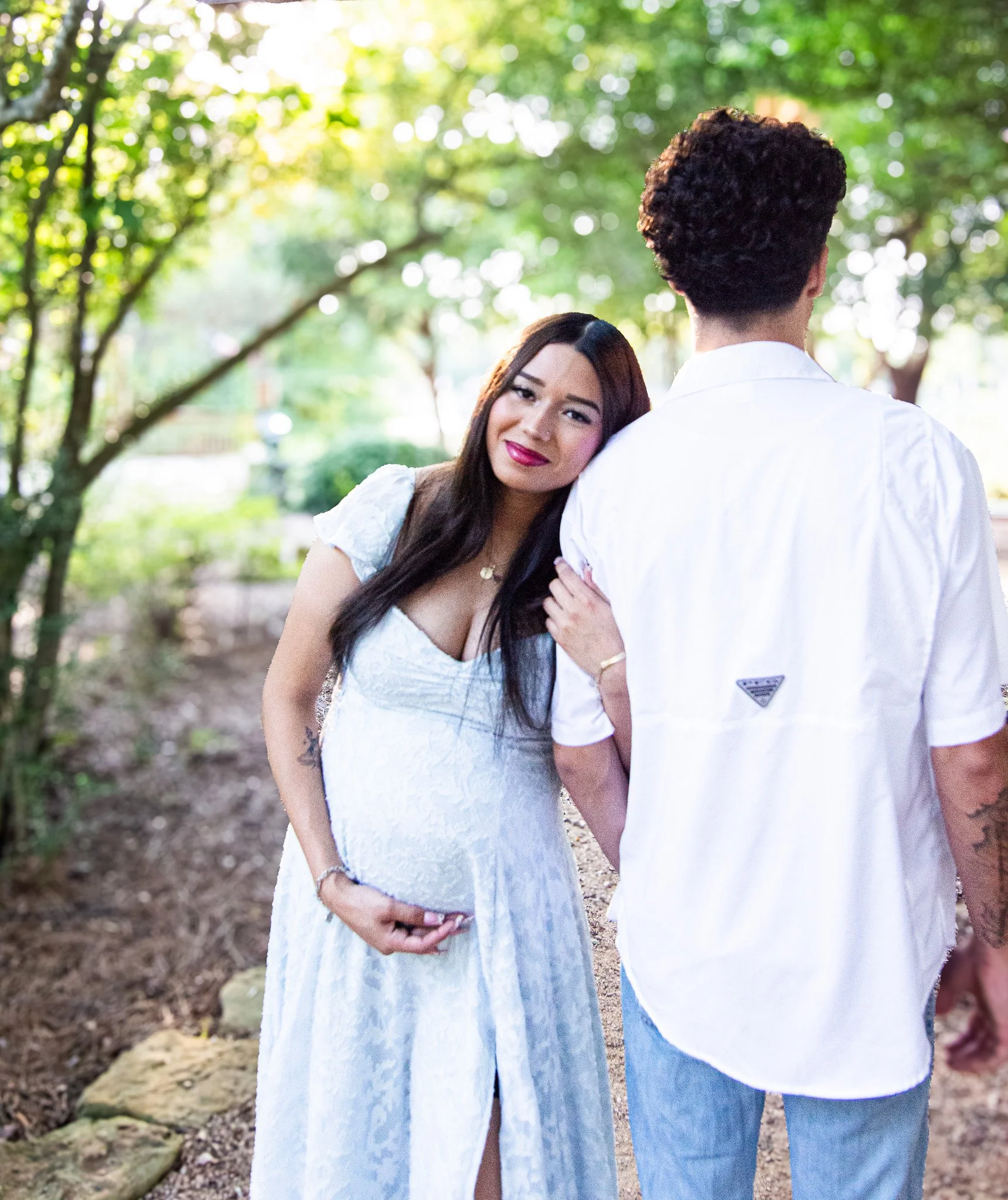 A pregnant woman in a white dress with long dark hair smiling and leaning on the shoulder of a man in a white shirt with curly dark hair, in a park with green trees and sunlight.