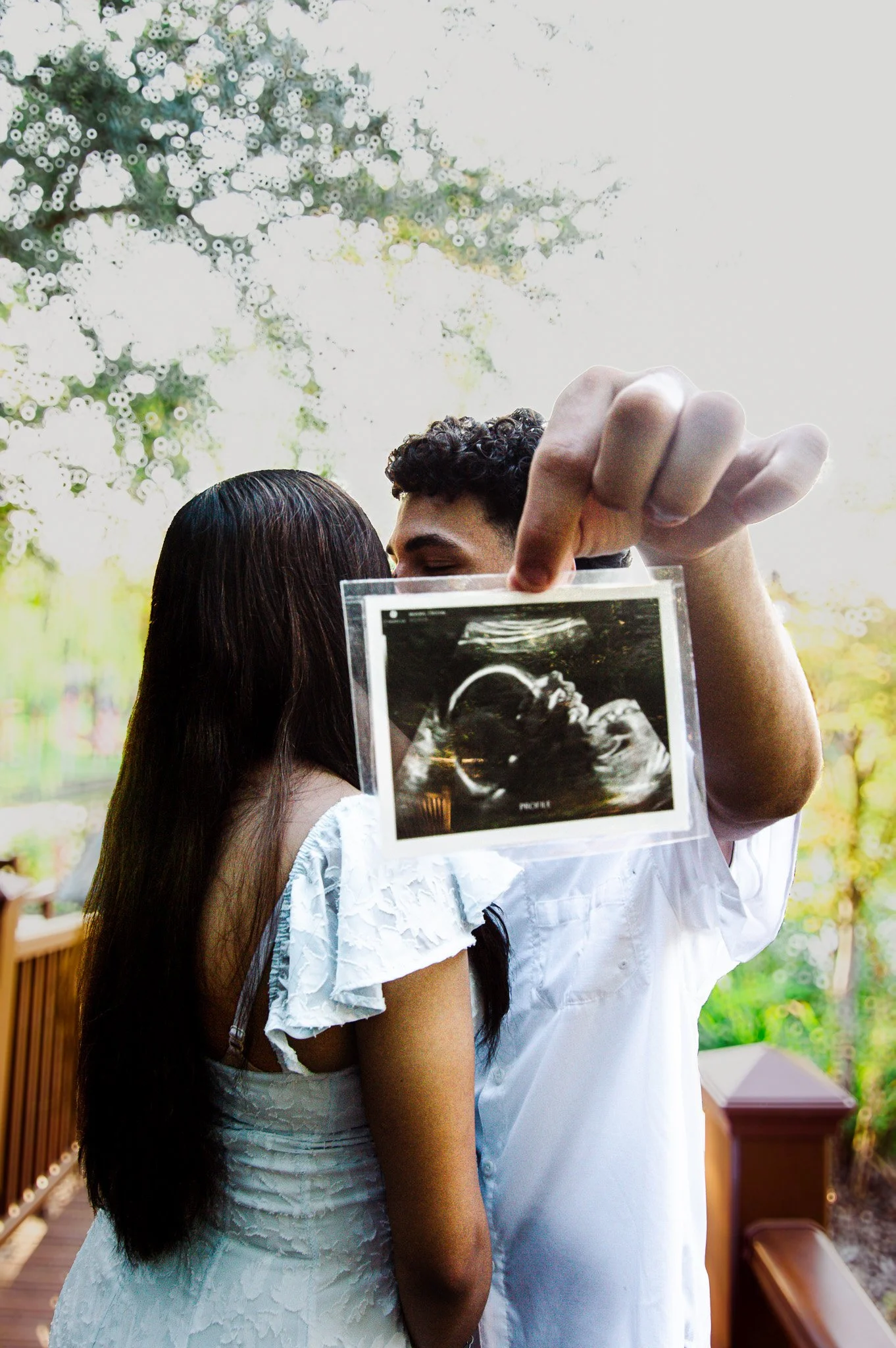A couple is embracing outdoors, with the man holding up an ultrasound picture of a baby in front of them.