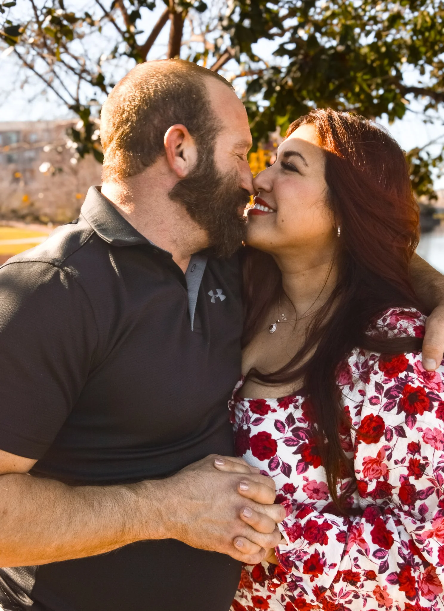 A man and woman embrace and smile at each other outdoors in a park, with trees and a building in the background.