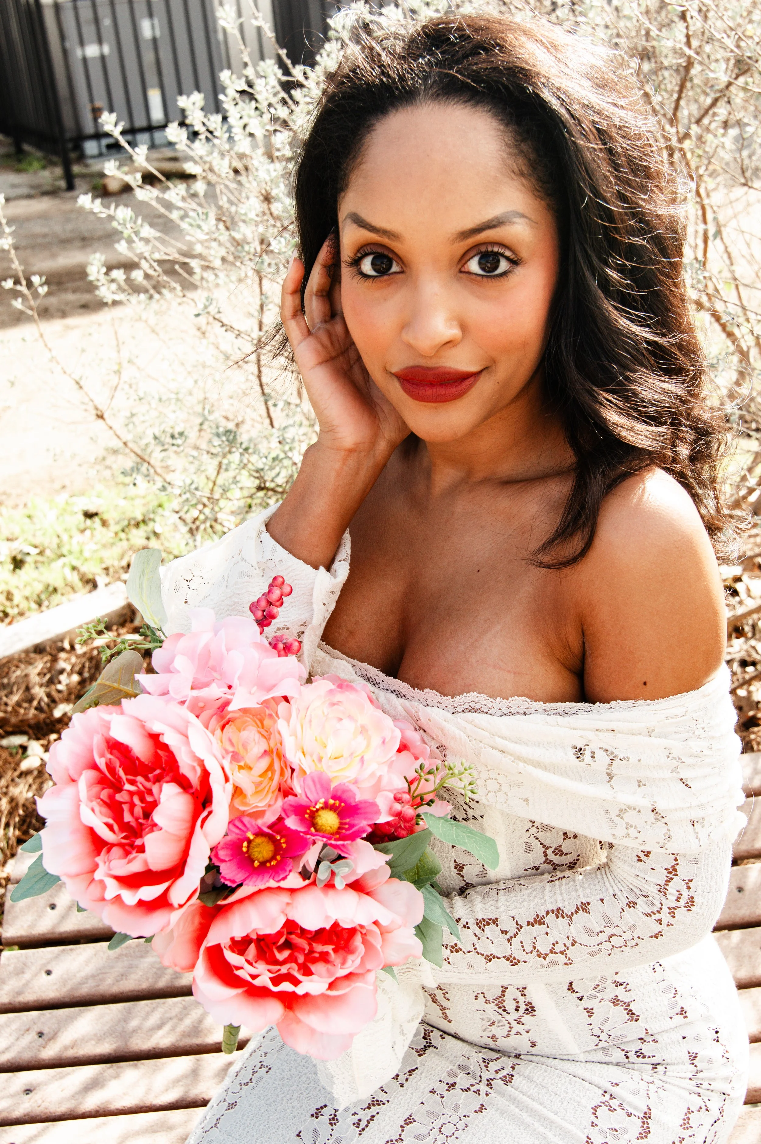 A woman with dark curly hair and red lipstick holding a pink and peach bouquet of flowers outdoors.