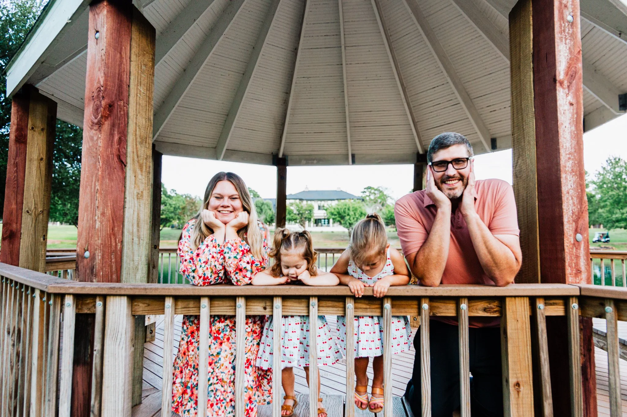 A family of four, with two young girls and two adults, leaning on a wooden railing inside a gazebo in a park, smiling at the camera.