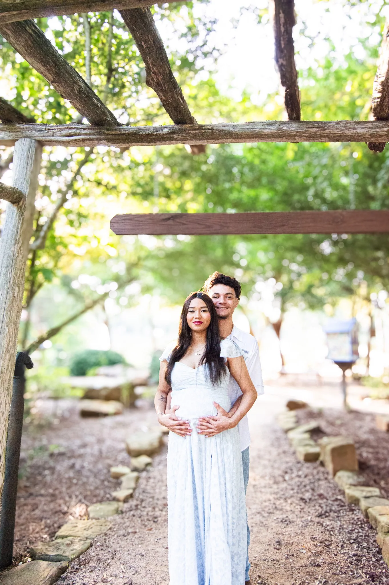 A pregnant woman and a man stand together outdoors under a wooden crossing, with trees and greenery in the background.
