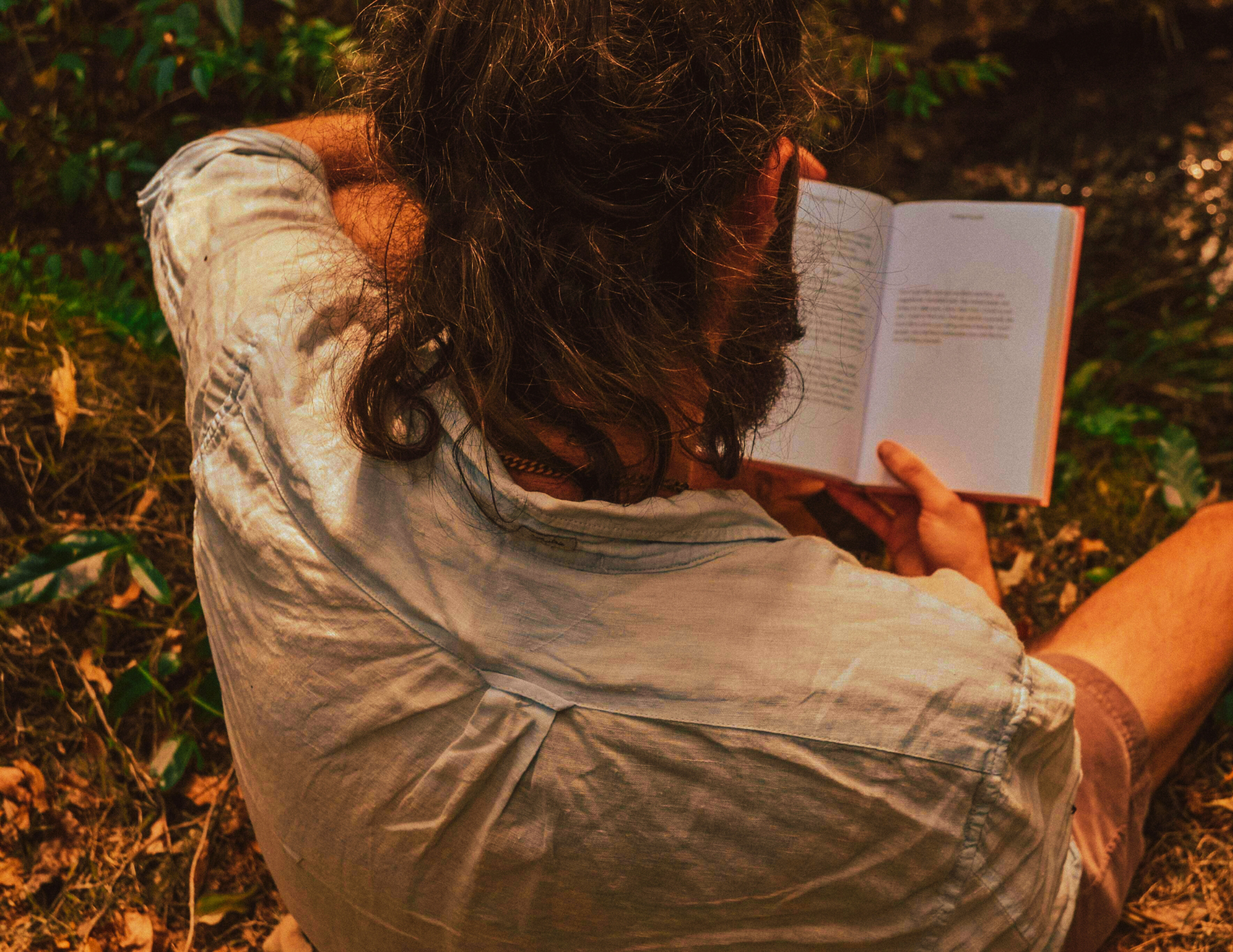 A man with long curly hair and a beard sitting outdoors on the ground, reading a book with a light-colored cover.