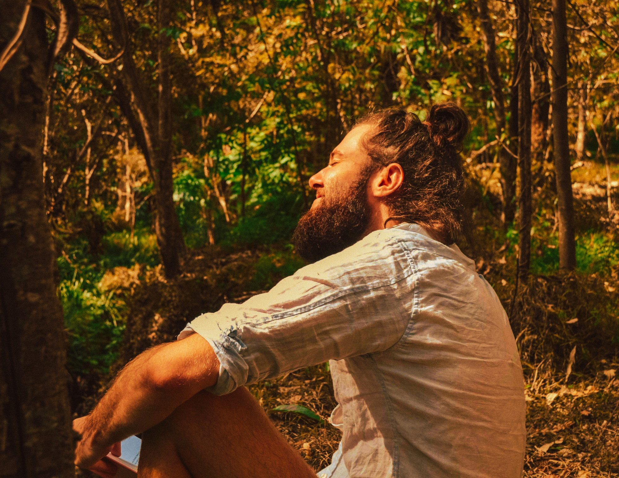 A man with a beard and long hair in a bun, wearing a light-colored shirt with rolled-up sleeves, sitting outdoors in a forest with closed eyes and a peaceful expression.