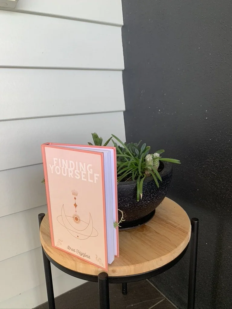 A small wooden table with black metal legs holding a black pot with green houseplants and a pink book titled 'Finding Yourself' by Brad Highfield.