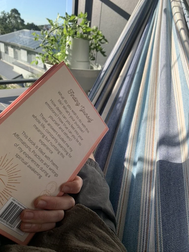 Person sitting on a striped hammock, holding a book titled 'Finding Yourself', with a potted plant and a view of neighboring houses on a sunny balcony in the background.