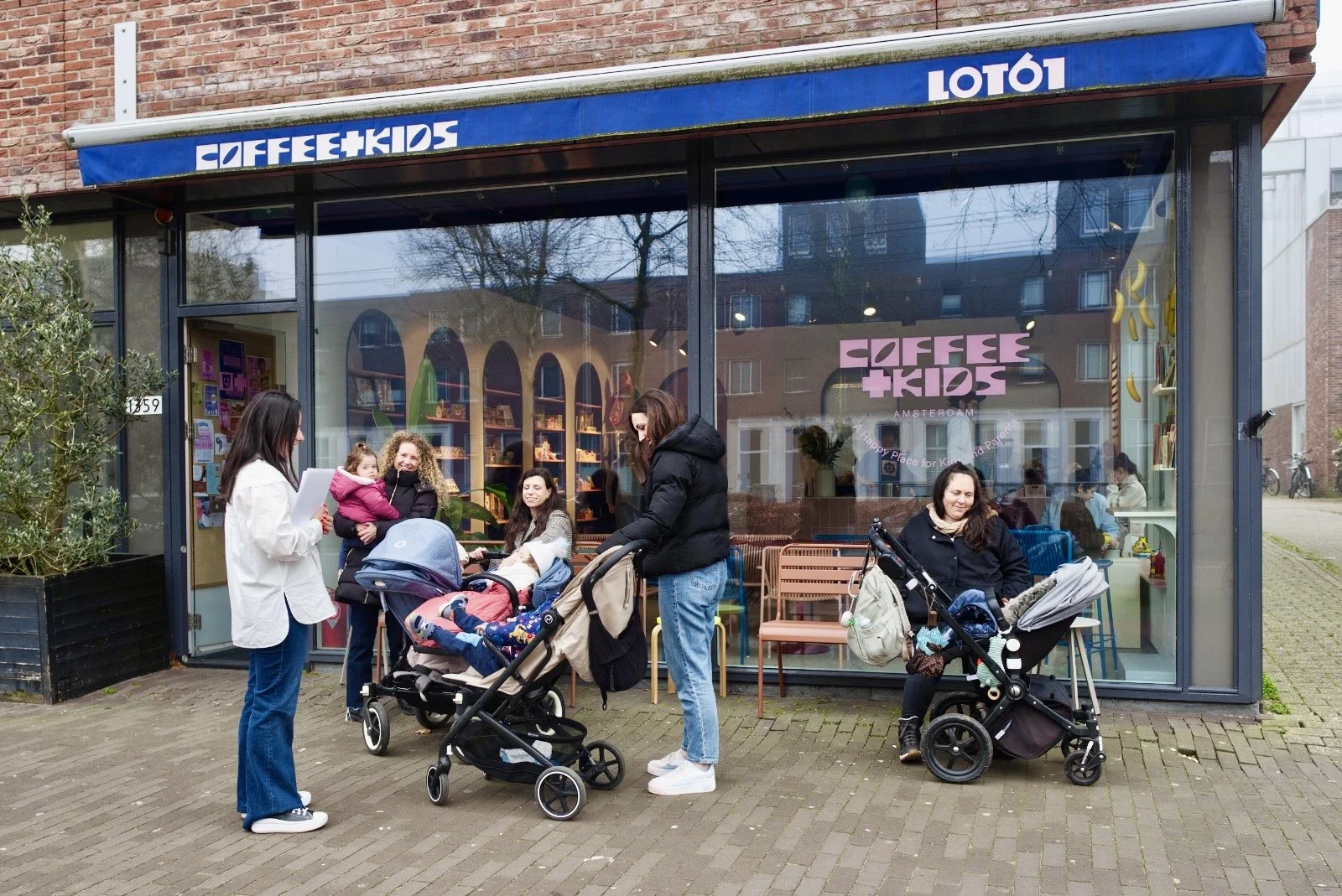 Group of women and children with strollers gathered outside a cafe called 'Coffee + Kids', with a glass storefront and interior filled with colorful decorations and books.