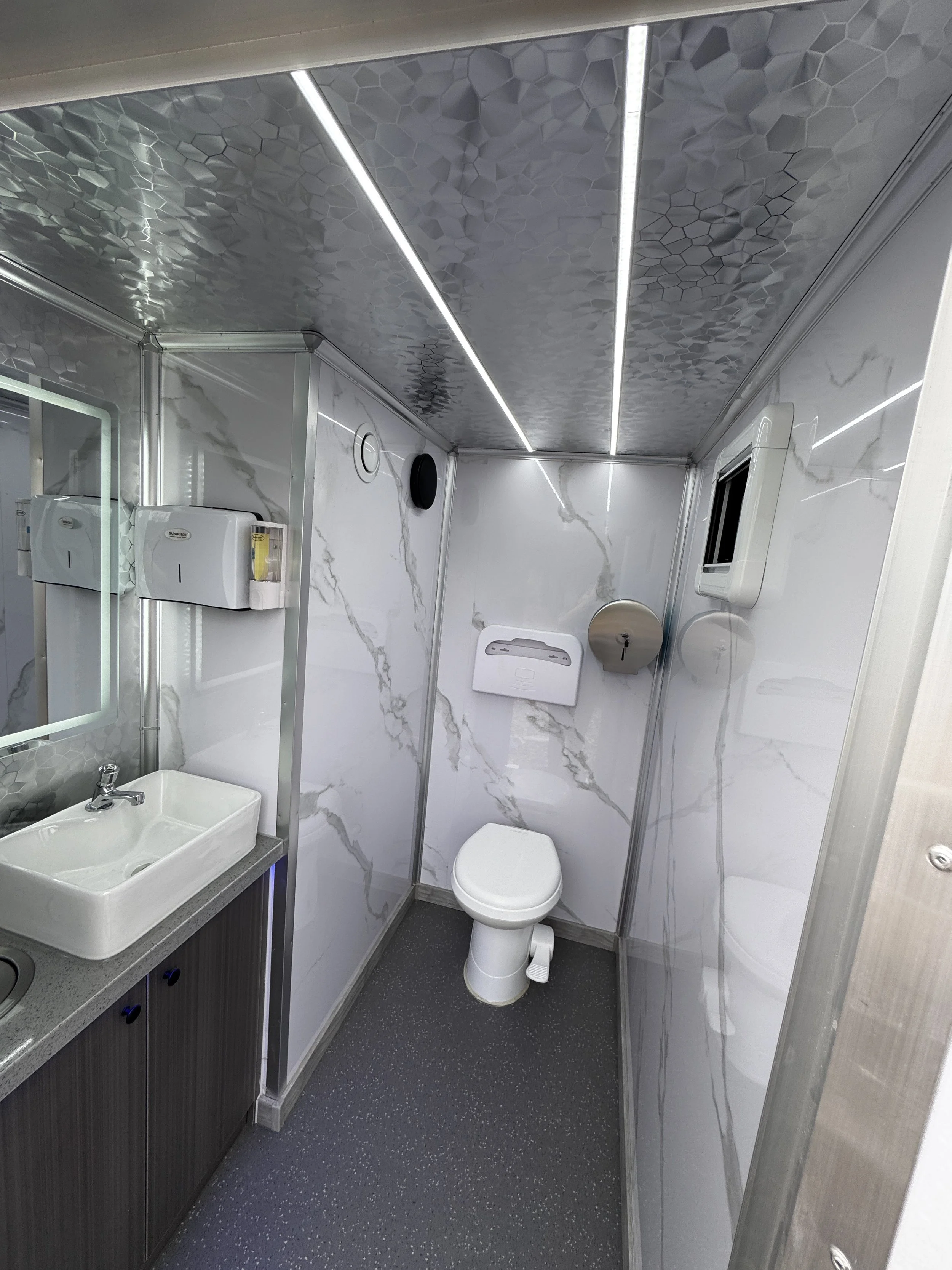 Small, modern restroom with white marble walls, a toilet, a sink with a mirror, and paper towel dispensers.