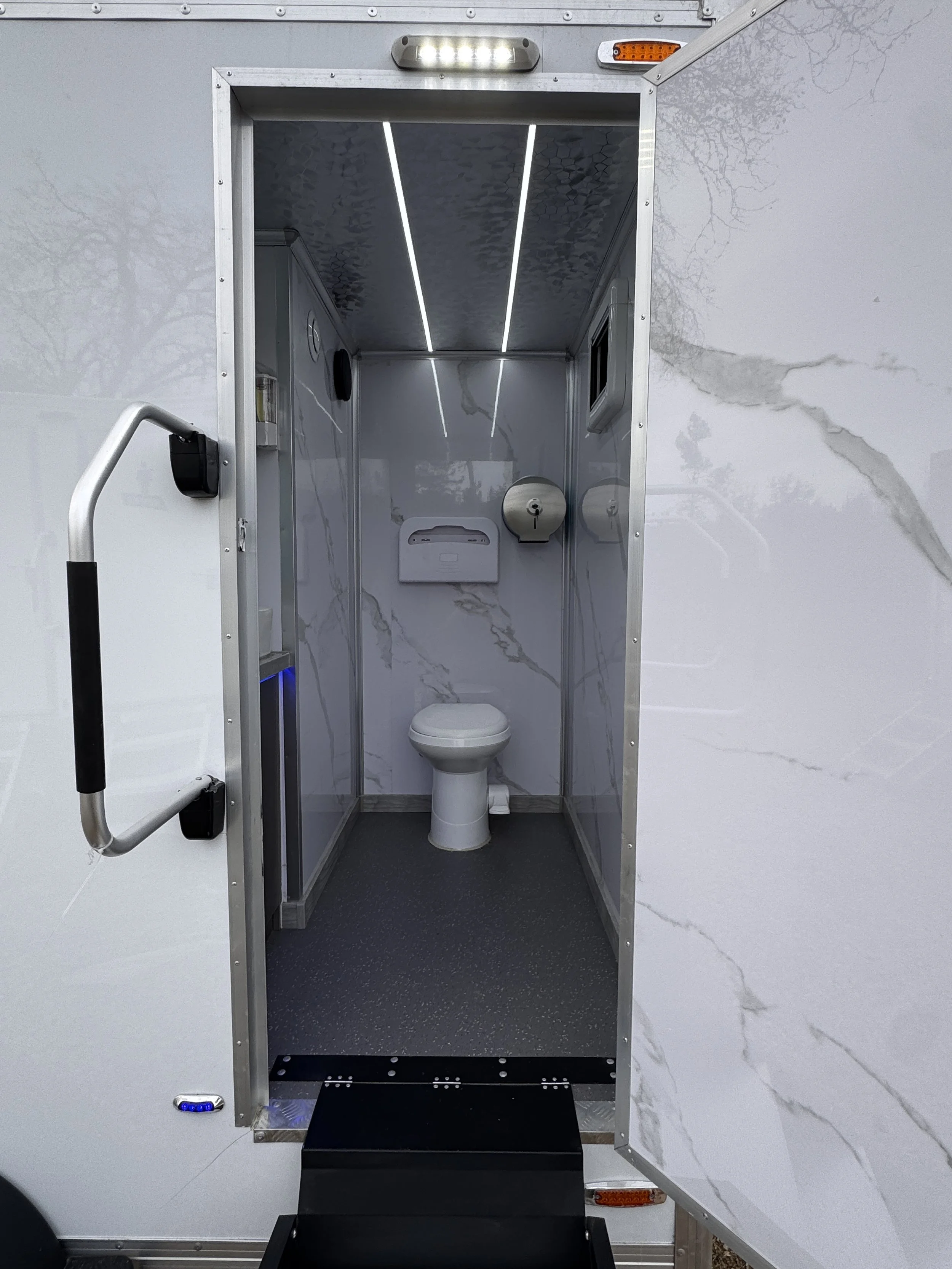 A compact mobile restroom with marble-like interior walls, a toilet, a paper towel dispenser, and small windows. It features LED lighting and handrails for accessibility.