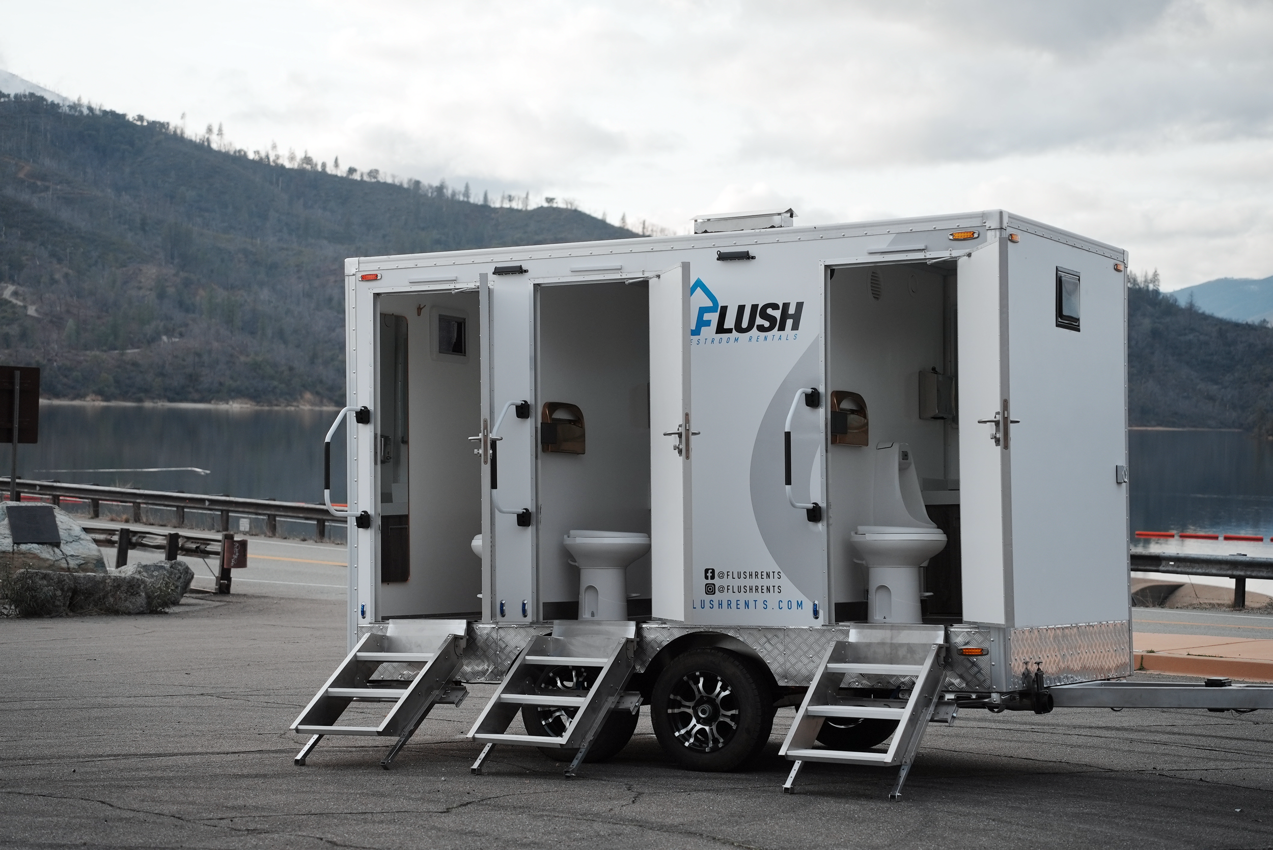 Mobile restroom trailer with open doors showing toilets inside, parked in a scenic outdoor location with mountains and water in the background.